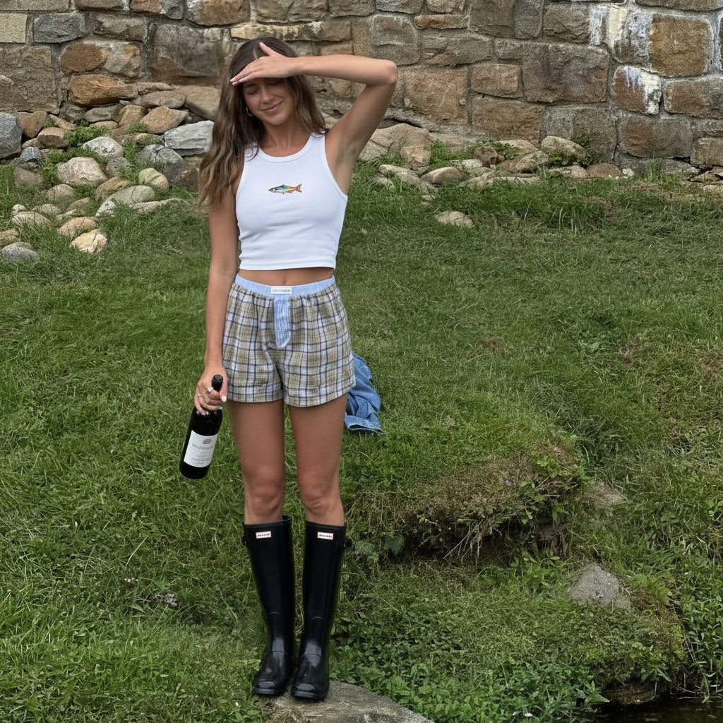 Woman in plaid shorts and black rain boots standing on grass with a stone wall in the background